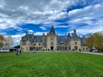 A grand historic estate with intricate architectural features and a large, manicured lawn in the foreground. The sky is partly cloudy with patches of blue peeking through. People are scattered across the lawn, and a tour bus is parked to the side.