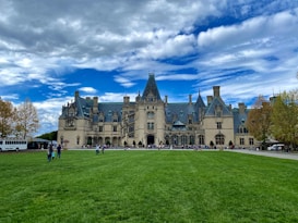 A grand historic estate with intricate architectural features and a large, manicured lawn in the foreground. The sky is partly cloudy with patches of blue peeking through. People are scattered across the lawn, and a tour bus is parked to the side.