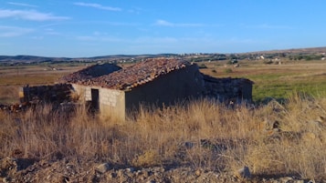 An old, rustic stone house with a tiled roof stands amidst a dry, grassy field. The structure appears weathered and abandoned, set against a backdrop of rolling hills and open skies. Sparse vegetation surrounds the building, highlighting its isolated setting.