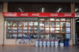 An outdoor view of a beverage shop with large window panes displaying various bottles. Above the window, there is a red Coca-Cola sign. In front of the shop, a blue bicycle is parked alongside a row of gas cylinders.