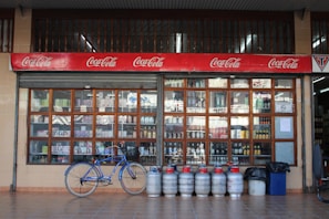 An outdoor view of a beverage shop with large window panes displaying various bottles. Above the window, there is a red Coca-Cola sign. In front of the shop, a blue bicycle is parked alongside a row of gas cylinders.
