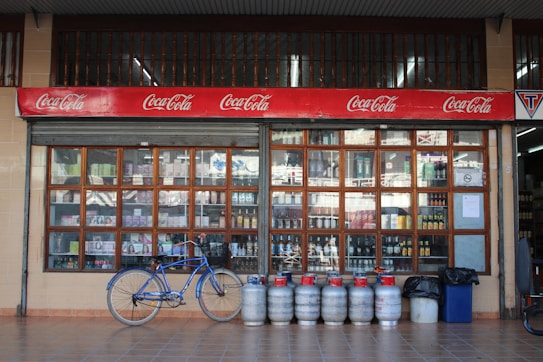 An outdoor view of a beverage shop with large window panes displaying various bottles. Above the window, there is a red Coca-Cola sign. In front of the shop, a blue bicycle is parked alongside a row of gas cylinders.