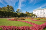 A beautifully landscaped garden featuring vibrant flower beds with pink and yellow flowers arranged in intricate patterns. Well-manicured green lawns and tall hedge rows provide a lush backdrop. A classical building with a light-colored facade and green roof is visible on the right, set against a blue sky with wispy clouds.