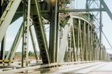 A large steel structure forms part of a bridge with intricate beams and trusses. The perspective provides a close-up view of the metalwork, emphasizing the geometric patterns. The bridge spans across a river or road, with a clear blue sky and some greenery visible in the background.