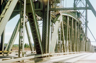 A large steel structure forms part of a bridge with intricate beams and trusses. The perspective provides a close-up view of the metalwork, emphasizing the geometric patterns. The bridge spans across a river or road, with a clear blue sky and some greenery visible in the background.