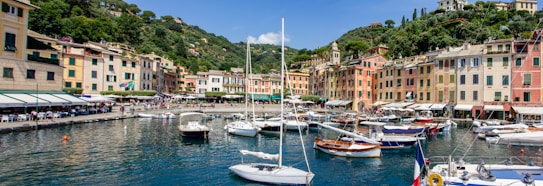 A picturesque seaside village with colorful buildings lining the waterfront. Numerous boats are docked in the clear blue harbor, surrounded by lush green hills. The scene is bustling with people enjoying the harbor area, and an Italian flag is visible.