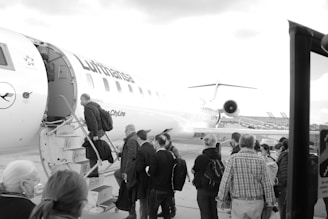 Passengers are boarding a Lufthansa airplane via a stairway. The image captures a line of people waiting to enter the aircraft. The plane is parked on an airport tarmac under a cloudy sky.