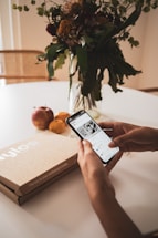 A person holding a smartphone displaying an app, with a cardboard box labeled 'Nylos' on a table alongside apples and an orange. In the background, there is a glass vase with a bouquet of assorted flowers and greenery.