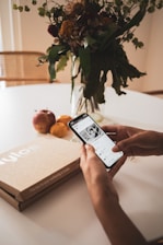 A person holding a smartphone displaying an app, with a cardboard box labeled 'Nylos' on a table alongside apples and an orange. In the background, there is a glass vase with a bouquet of assorted flowers and greenery.