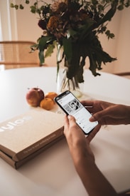 A person holding a smartphone displaying an app, with a cardboard box labeled 'Nylos' on a table alongside apples and an orange. In the background, there is a glass vase with a bouquet of assorted flowers and greenery.