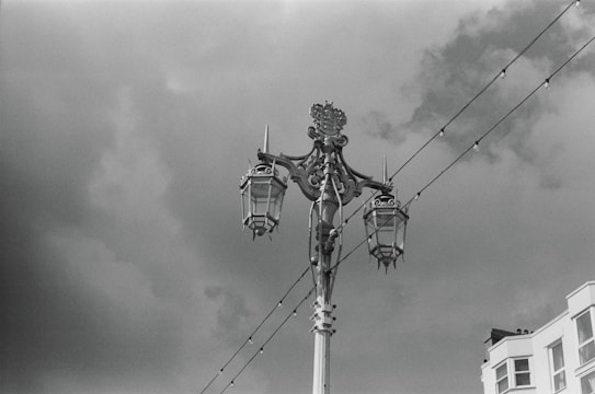 A decorative street lamp post with ornate detailing is set against a cloudy sky. Two lanterns hang on either side of the central post, which features intricate metalwork. In the background, strings of lights extend horizontally above, and a building with white facades is partially visible on the right.