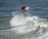 A surfer performs an aerial maneuver above a breaking wave, showcasing skill and balance. The ocean water is turbulent, with white foam and spray around the surfer.