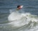 A surfer performs an aerial maneuver above a breaking wave, showcasing skill and balance. The ocean water is turbulent, with white foam and spray around the surfer.