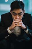 A person in a formal black suit sits with hands clasped closely in front of the face, looking directly at the camera. The setting features a blurred blue-toned background with soft lighting that adds a contemplative mood.