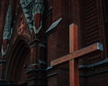 An ornate brick church facade with detailed carvings and gothic arches. A large wooden cross is prominently placed in front of the church, inscribed with Cyrillic text.