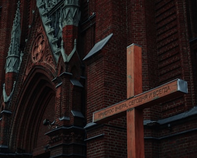 An ornate brick church facade with detailed carvings and gothic arches. A large wooden cross is prominently placed in front of the church, inscribed with Cyrillic text.