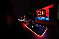 A dark gaming setup featuring a glowing mouse, mouse pad, and keyboard with vivid RGB lighting. The open computer case reveals components illuminated in red and blue lights, accentuating the high-tech atmosphere.