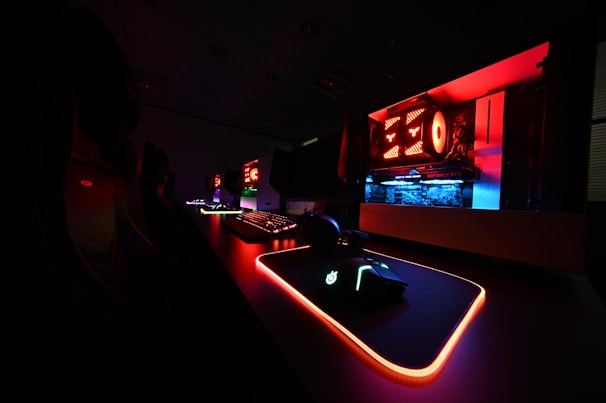A dark gaming setup featuring a glowing mouse, mouse pad, and keyboard with vivid RGB lighting. The open computer case reveals components illuminated in red and blue lights, accentuating the high-tech atmosphere.