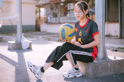 A person sitting on a concrete slab in an outdoor area, holding a blue and yellow volleyball. The individual is wearing sports attire, including a black shirt with red accents and black pants with white stripes. Glasses are worn, and the setting appears to be a sports court with metal structures in the background. Sunlight casts shadows on the ground.