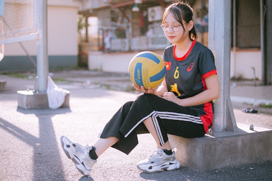 A person sitting on a concrete slab in an outdoor area, holding a blue and yellow volleyball. The individual is wearing sports attire, including a black shirt with red accents and black pants with white stripes. Glasses are worn, and the setting appears to be a sports court with metal structures in the background. Sunlight casts shadows on the ground.