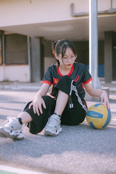 A person with glasses is sitting on the ground in a casual manner, holding a yellow and blue volleyball. They are wearing a dark sports jersey with red accents, black pants, and sneakers. The background shows an exterior setting with a building, featuring windows and a metal pole.