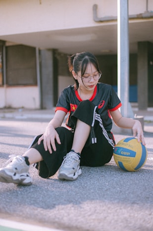 A person with glasses is sitting on the ground in a casual manner, holding a yellow and blue volleyball. They are wearing a dark sports jersey with red accents, black pants, and sneakers. The background shows an exterior setting with a building, featuring windows and a metal pole.