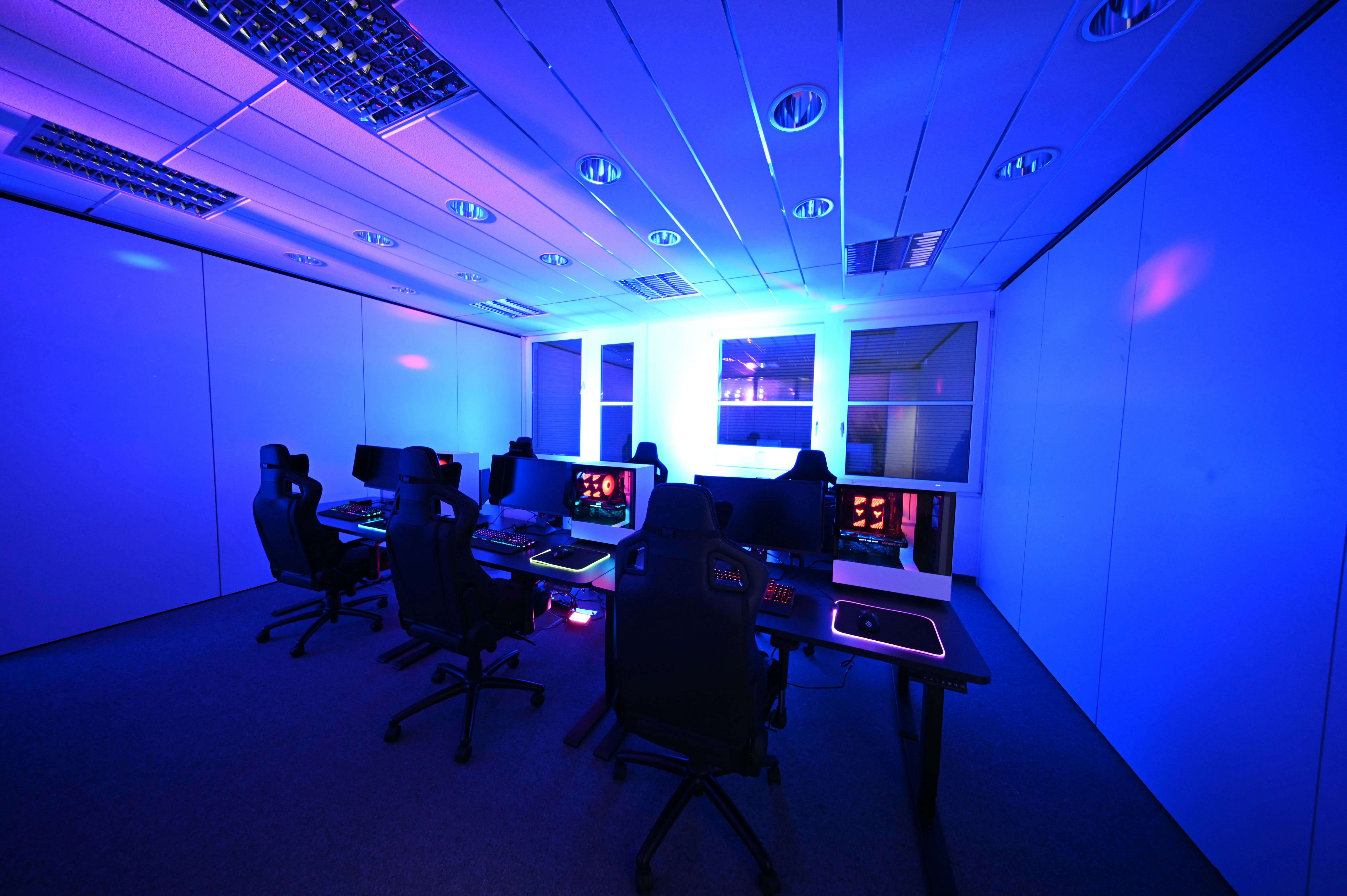 A modern gaming setup with multiple high-backed gaming chairs and monitors arranged in a room with a cool blue lighting. The computers have illuminated keyboards and glowing screens, creating a high-tech ambiance.