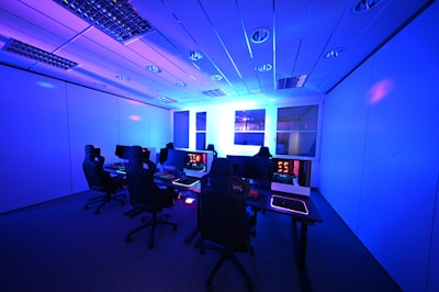 A modern gaming setup with multiple high-backed gaming chairs and monitors arranged in a room with a cool blue lighting. The computers have illuminated keyboards and glowing screens, creating a high-tech ambiance.