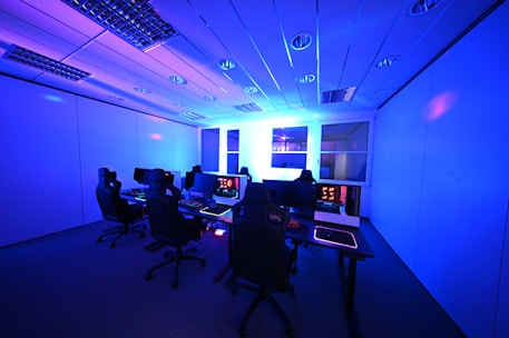 A modern gaming setup with multiple high-backed gaming chairs and monitors arranged in a room with a cool blue lighting. The computers have illuminated keyboards and glowing screens, creating a high-tech ambiance.