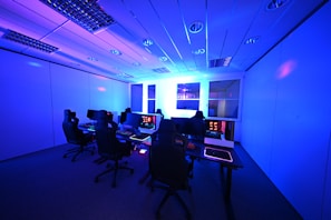 A modern gaming setup with multiple high-backed gaming chairs and monitors arranged in a room with a cool blue lighting. The computers have illuminated keyboards and glowing screens, creating a high-tech ambiance.