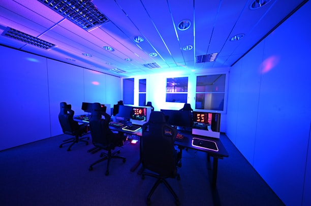 A modern gaming setup with multiple high-backed gaming chairs and monitors arranged in a room with a cool blue lighting. The computers have illuminated keyboards and glowing screens, creating a high-tech ambiance.