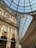 A grand, ornately decorated shopping arcade with an arched glass ceiling. The architecture features intricate details and classical elements. Elegant shop fronts can be seen, including a Prada store. The light filtering through the glass ceiling creates a bright, inviting atmosphere.