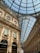 A grand, ornately decorated shopping arcade with an arched glass ceiling. The architecture features intricate details and classical elements. Elegant shop fronts can be seen, including a Prada store. The light filtering through the glass ceiling creates a bright, inviting atmosphere.