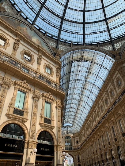 A grand, ornately decorated shopping arcade with an arched glass ceiling. The architecture features intricate details and classical elements. Elegant shop fronts can be seen, including a Prada store. The light filtering through the glass ceiling creates a bright, inviting atmosphere.
