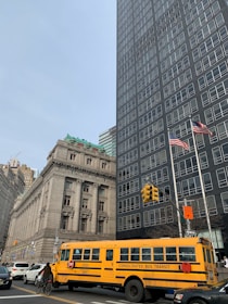 A yellow school bus with the words 'Consolidated Bus Transit' is moving through a city intersection. There are two American flags on flagpoles situated next to a tall, dark glass building. Another historical building with ornate architecture is visible in the background. Several vehicles and a cyclist can be seen on the road.