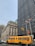 A yellow school bus with the words 'Consolidated Bus Transit' is moving through a city intersection. There are two American flags on flagpoles situated next to a tall, dark glass building. Another historical building with ornate architecture is visible in the background. Several vehicles and a cyclist can be seen on the road.
