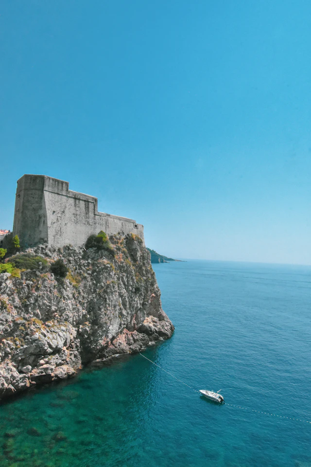 Fort Lovrijenac perched on a rocky sea cliff, with a boat anchored in the turquoise water below