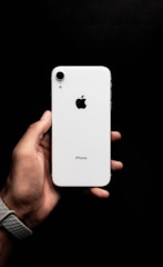 A hand holding a white smartphone with a visible brand logo and camera on the back. The background is a solid black, enhancing the contrast with the phone.
