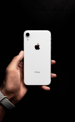 A hand holding a white smartphone with a visible brand logo and camera on the back. The background is a solid black, enhancing the contrast with the phone.