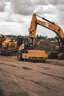 Heavy construction machinery, including a large excavator and a bulldozer, are operating on a dirt construction site. The ground shows tracks from the machinery, and the sky above is overcast with clouds.