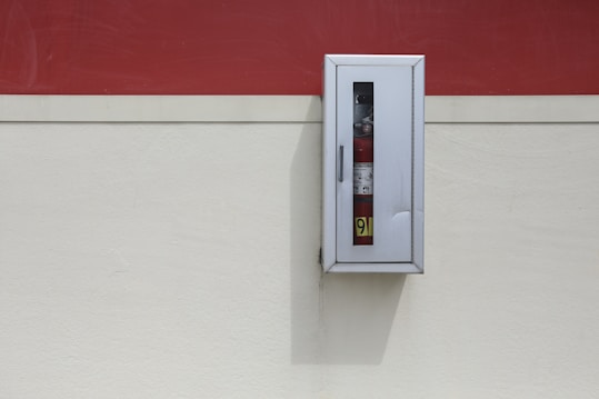 A fire extinguisher is encased within a metal cabinet mounted on a wall. The background consists of two-tone colors, with a red section at the top and a cream section at the bottom. The cabinet casts a shadow on the wall.