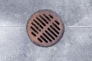 A circular metallic drain cover rests on a weathered concrete surface. The cover is rusty, showing signs of age and exposure to the elements. Uniform grid slots allow water drainage.