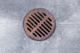 A circular metallic drain cover rests on a weathered concrete surface. The cover is rusty, showing signs of age and exposure to the elements. Uniform grid slots allow water drainage.