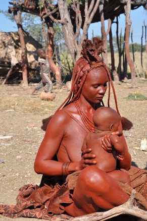 A woman with distinctive hair and skin treatments sits outdoors, holding a baby in her arms. The background shows a structure made of wooden poles and another child walking nearby. The setting appears to be rural and earthy.
