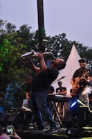 An enthusiastic adult playing the saxophone outdoors with leafy trees behind.