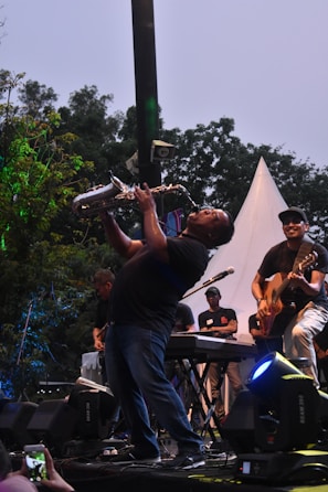 An enthusiastic adult playing the saxophone outdoors with leafy trees behind.