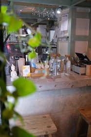 A cozy cafe setting featuring a countertop with glass bottles, a credit card terminal, and a small statue. There are wine glasses hanging upside down above, and some green leaves in the foreground, creating a natural feel.