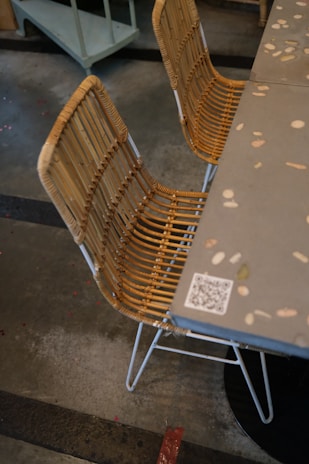 A rattan chair with a metal base is positioned next to a gray table decorated with various small embedded colored stones. The floor is concrete with visible markings, and a QR code is visible on the table's edge.