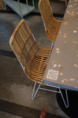 A rattan chair with a metal base is positioned next to a gray table decorated with various small embedded colored stones. The floor is concrete with visible markings, and a QR code is visible on the table's edge.