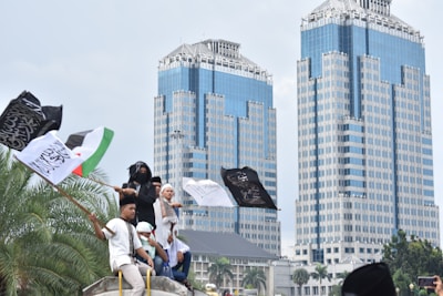 A group of people stand on a vehicle holding several flags with Arabic text, set against the backdrop of two modern skyscrapers. Palm trees are visible in the foreground, and the mood suggests a sense of demonstration or gathering.